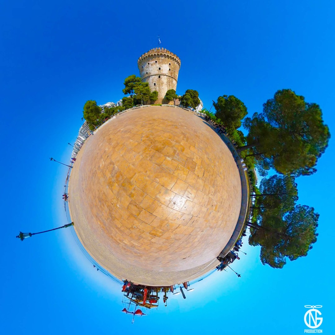 Day tiny-planet 360° view of the White Tower with a sunlit promenade and Aegean-blue sky.